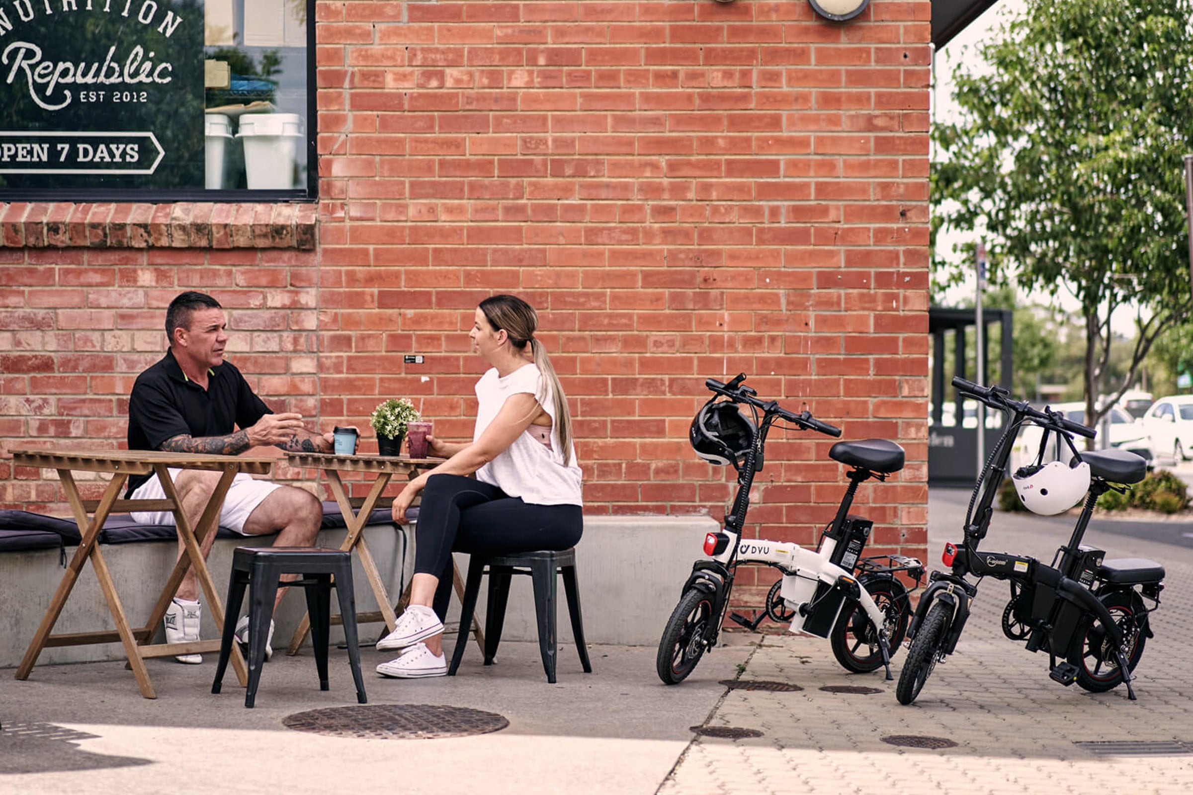 Twee DYU elektrische fietsen T1 en A5 staan naast een jonge vrouw en man in een caféomgeving.